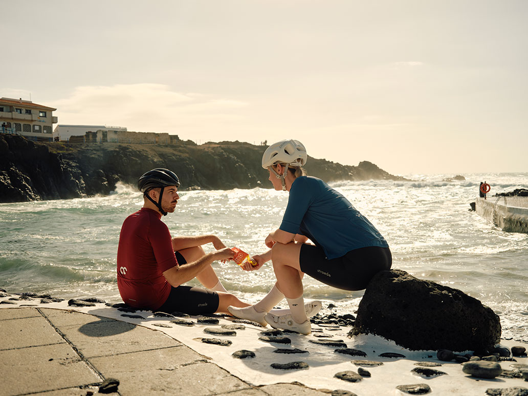 Zwei Radfahrer in minimalistischer Radbekleidung sitzen und essen am Meer